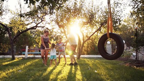 Children having fun holding hands with parents at park with tire swing in foreground and sunlight coming through trees. Having joined hands are turned around