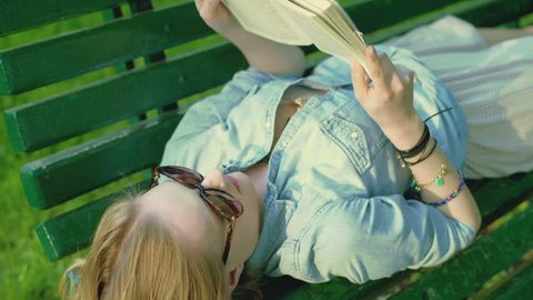 Pretty girl lying on the wooden bench and reading book, steadycam shot
