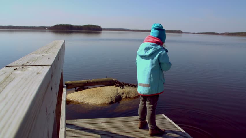 Little girl plays at coast of the lake. She throws the small stones in Water.