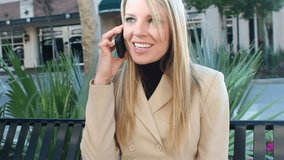A pretty businesswoman on a cell phone sits down on a sidewalk bench