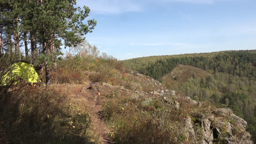 Young blonde woman tourist comes out of camping tent, goes to the cliff,  looking  on the landscape and stretching with pleasure. Russia, Siberia, Salair