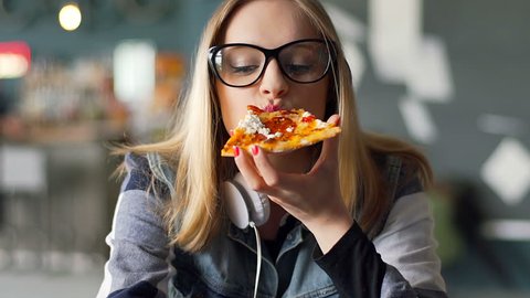 Hipster girl eating pizza in the restaurant and smiling to the camera
