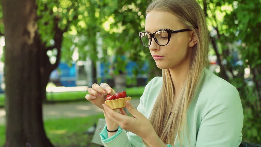 Pretty girl sitting on the bench in the park and eating a cookie
