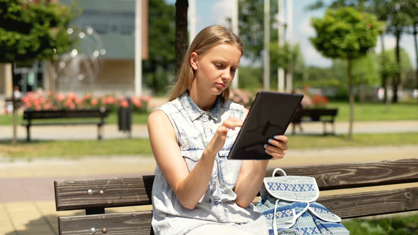 Pretty student sitting on the bench and browsing internet on tablet
