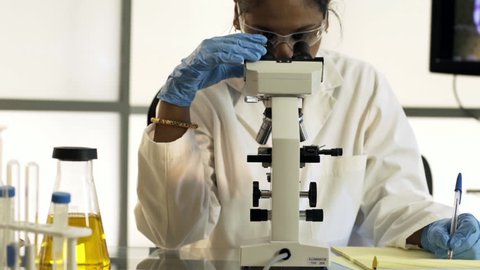 A female chemist of Indian ethnicity working in a lab documenting her observations made through a microscope.