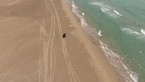Aerial shooting from flying drone of a woman tourist is riding on a rental ATV transport on sandy beach near sea with turquoise water. Young hipster female having extreme sport leisure with quad bike
