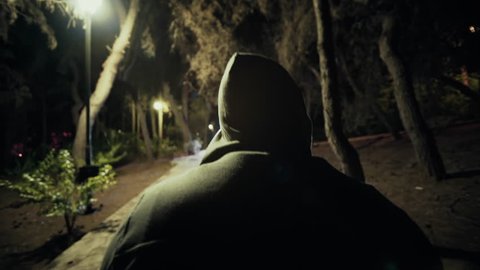 Suspicious hooded figure walks in a dark park at night,gimbal tracking.A suspicious man wearing a hoodie walks in a dark ominous urban park at night
