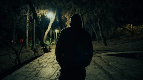 Suspicious hooded figure walks in a dark park at night,gimbal tracking.A suspicious man wearing a hoodie walks in a dark ominous urban park at night