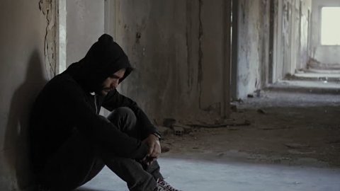 Hooded young man inside destroyed abandoned building,back against wall,slow motion, dramatic.Young man with social issues, inside a big demoloshed building in 100fps.Camera gimbal or crane motion.