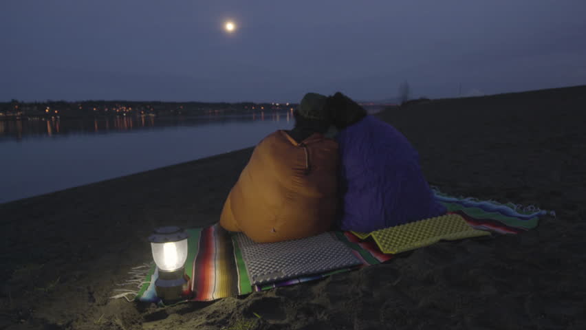 Couple Sit On A Riverbank, Wrapped In Sleeping Bags, Enjoy The View Of River