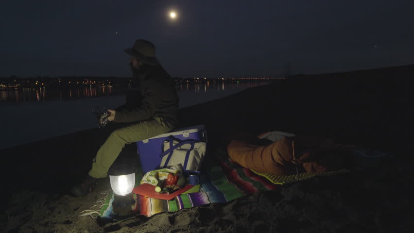 Man Sits On His Cooler, Plays Guitar And Sings In The Moonlight On Sandy Riverbank