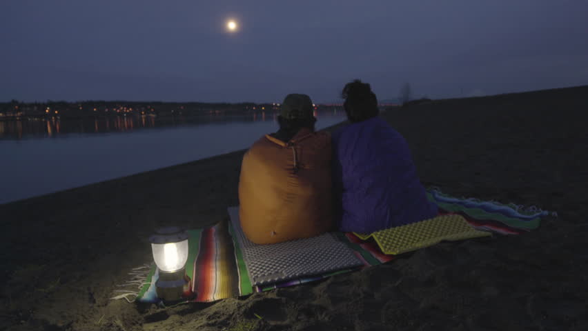 Couple Sit And Talk, Wrapped In Sleeping Bags, Enjoy View Of Moon Over Riverbank