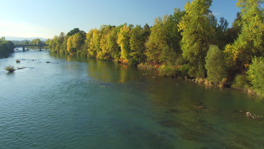 AERIAL: Flying above wide fast current river with lush bushy riverbank running towards big urban city and speedway bridge. Big town and high skyscrapers in the distance with mountains in background