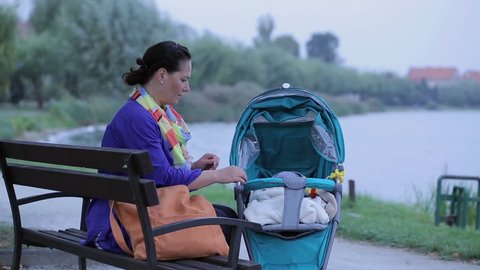 Mother is draw out a baby from stroller, sitting on bench near lake
