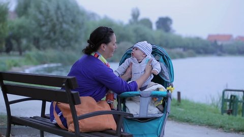Mother is draw out a little son from stroller, sitting on bench 
