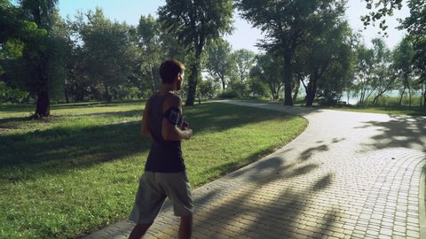 jogger jogging in the morning outdoors, rear back view. adult man using smartphone listen music wearing in t-shirt and sneakers running in the park, where walking man with dogs