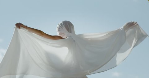 Dancer expressing herself with neo classical dance moves. Ballet trained dancer. Using a white satin sheet against the blue sky with water in the background. Shot in slow motion in an artistic way.