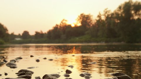 CLOSE UP, DOF: Stunning riverbank shore with big rocks and stones at magical sunrise. Glossy river surface reflecting beautiful fiery sun hiding behind tall lush trees and at magical golden sunset