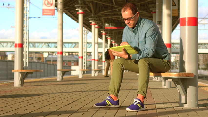 Bald nerdy man in glasses sitting at railroad station and using his tablet computer at sunset. 4K shot