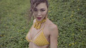 Closeup half body shot of beautiful brunette woman in yellow crochet bikini smiling and posing outside over green grass and flowers background - video in slow motion