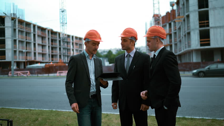 4K. ULTRA HD. Three youg successful Customers in orange helmets sign architect project on black tablet PC with newly constructed buildings at the background. Teal-and-orange middle shot.