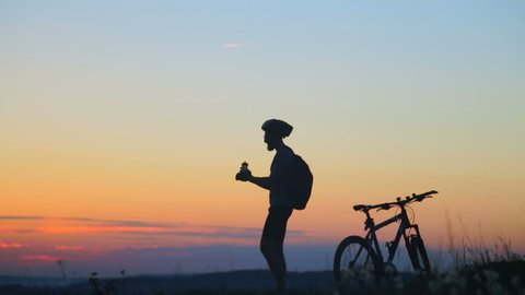 Silhouette bicyclist drinks water on top of a mountain. Slow motion.