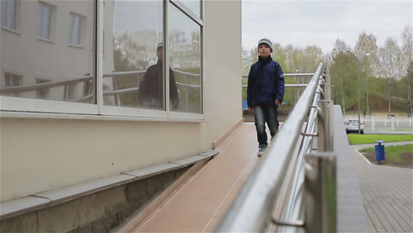 A small boy walking down a wheelchair ramp. Slow motion. It is a spring cloudy day. There is a man in the background walking and talking on the phone.