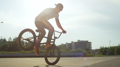 SLOW MOTION CLOSE UP: Extreme bmx biker riding in sunny park, stopping the bmx bike and doing nollie tail whip trick on beautiful summer day. Cool young bmx biker performing tricks on city street