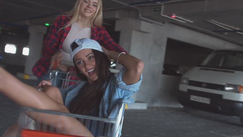 MED fashion funny young hipster girls having fun at the shopping mall parking, spinning each other in shopping cart. 60 FPS 4K UHD