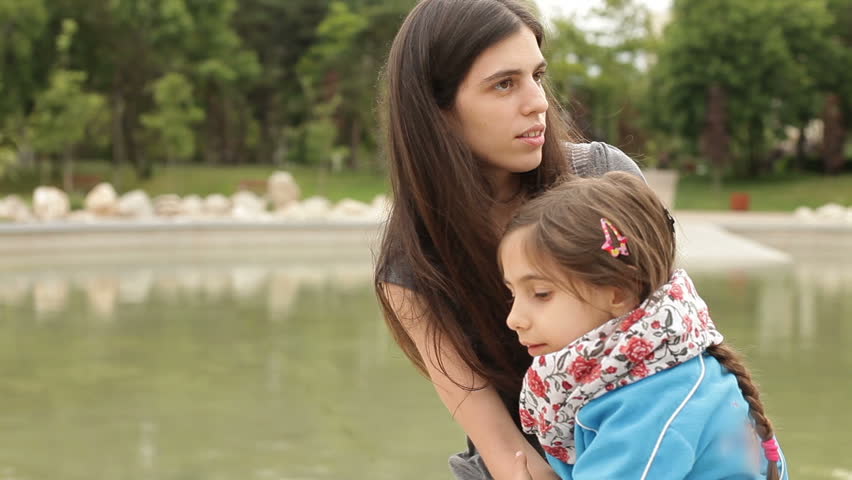 Mother and daughter hugging, having fun. Little gossip. Outdoor long shot, cloudy weather, beside lake, in park.