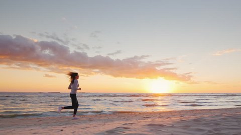 Young attractive woman jogging on the beach at sunset Full length. Beautiful clouds and breathtaking sunset. Steadicam slow motion video.