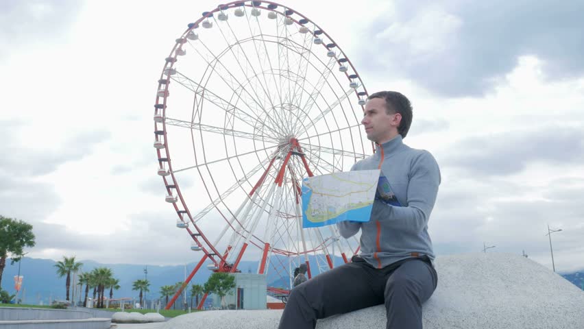 Young handsome man looking for landmarks on a paper map nearby ferris wheel. Early morning. The wheel does not spin.