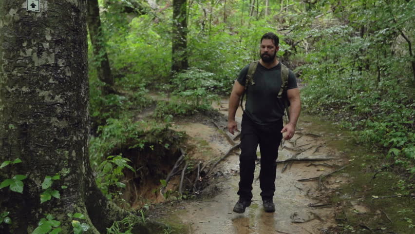 A male hiker walks beside a tree and a river along a path, in slow motion.