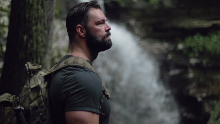 A male hiker stands near a waterfall de-focused in slow motion.