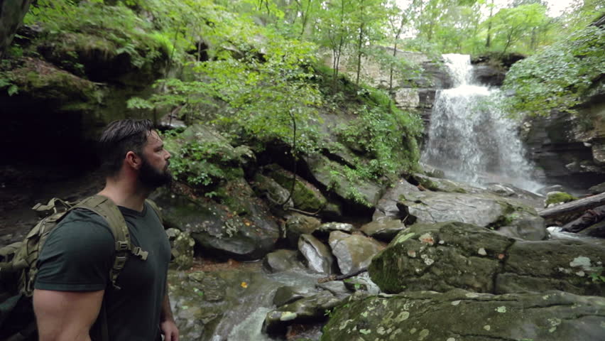 A male hiker stands at the bottom of a large waterfall in slow motion, shallow focus.