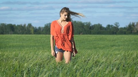 Full lenght portrait of a beautiful blonde young romantic woman in a red shirt walking slowly with a sexy look at the green field