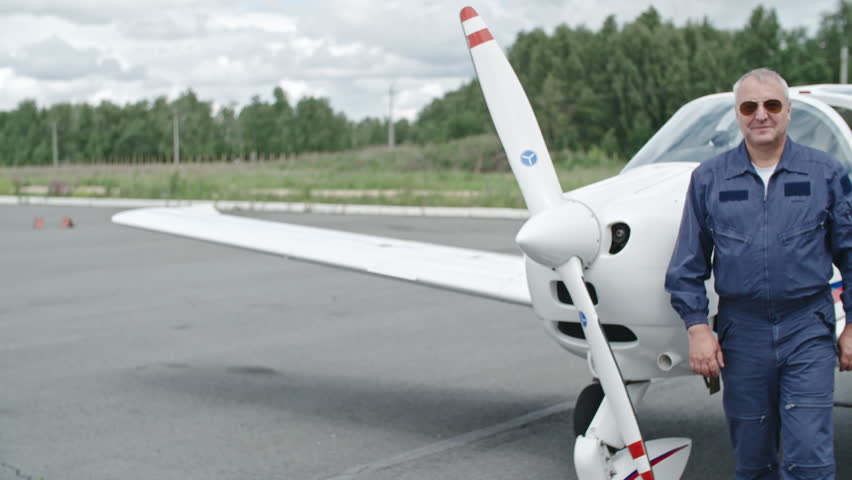 Mature man with aviator sunglasses standing with his arms crossed before small aircraft on airfield and looking at camera