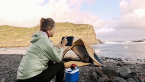 4k medium of Independent traveler woman using tablet device and using banking application next to yellow tent with camping gear on an epic camping adventure.