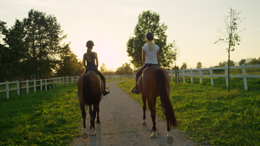 SLOW MOTION, CLOSE UP: Two young girls horseback riding amazing brown stallions walking along sandy footpath into magical golden sunset on big horse ranch farm. Friends on morning ride at sunrise