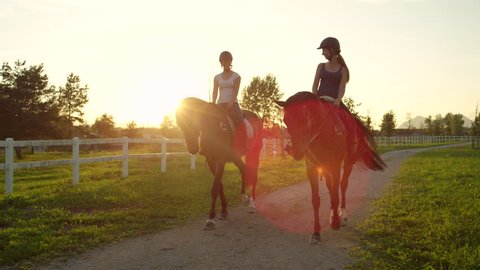 SLOW MOTION, CLOSE UP: Two cheerful girls horseback riding amazing strong brown horses walking along footpath at magical golden sunset on beautiful horse ranch farm. Friends on relaxing morning ride