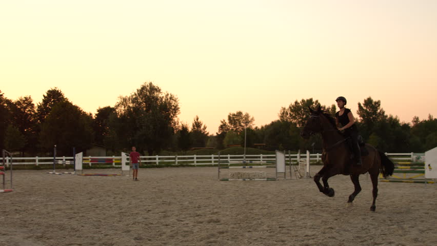 SLOW MOTION CLOSE UP: Young girl horseback riding strong brown horse jumping the fence in outdoors sandy parkour dressage arena at sunrise. Competitive rider training jumping over obstacles at sunset Royalty-Free Stock Footage #19799338
