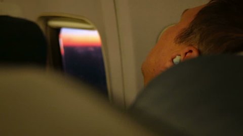 Young man resting on the plane during the flight. He sleeps in the headphones. The porthole beautiful sunrise.