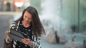 Happy young woman with tablet computer having video chat while sitting on the bench at the street, fountain background