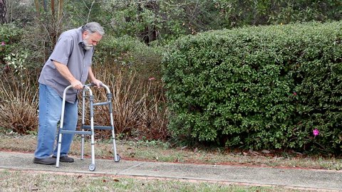 Disabled man uses a walker to support himself as he walks along a sidewalk.