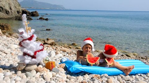 Kids in chrismas hat are eating watermelon on the beach