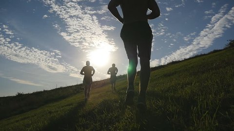 Young men running over green hill over blue sky background. Male athletes is jogging in nature at sunset. Sport runners jogging uphill outdoor at sunrise with flare. Cross-country training. Slowmotion