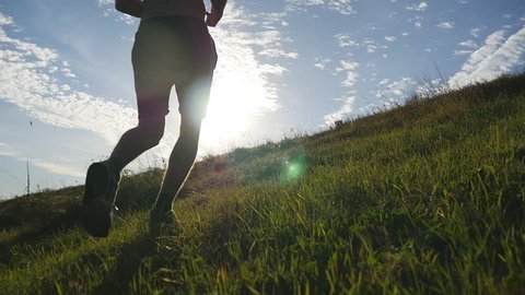 Young man running over green hill over blue sky background. Male athlete is jogging in nature at sunset. Sports runner jogging uphill outdoor at sunrise with flare. Cross-country training. Slow motion