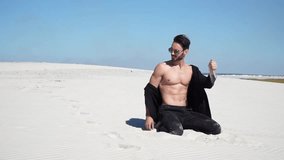 Man model appearance in a black jacket and trousers sitting on the sand