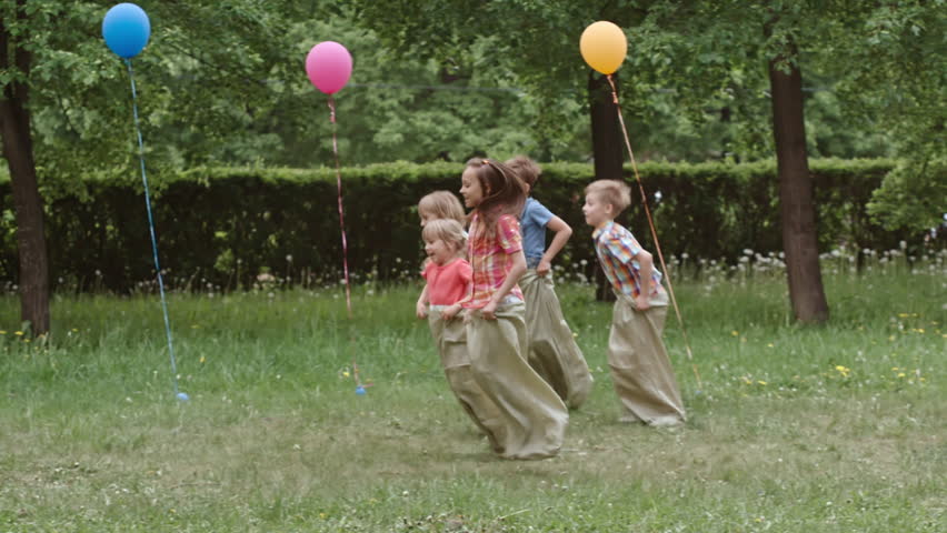 Side view of children jumping in sacks through the green lawn in the park while having birthday party with parents