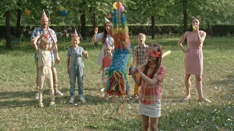 Slow motion shot of girl with covered eyes hitting pinata with wooden bat during outdoor birthday party with friends and parents tossing confetti and blowing party horn